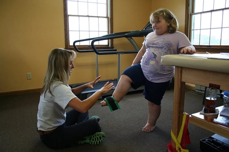 Physical therapist assisting a young child on a parallel bar exercise in a therapy room with yellow walls and windows.