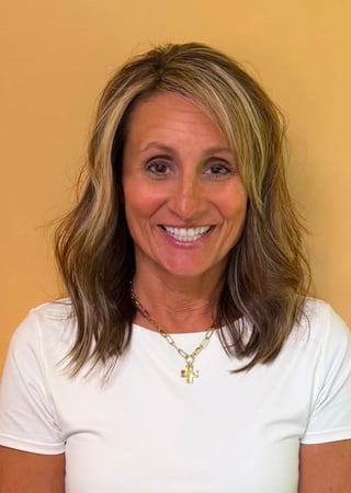 Portrait of a woman with shoulder-length brown hair wearing a white t-shirt and gold cross necklace, smiling against a yellow background