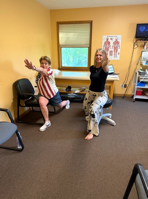 Two people performing a fitness movement in a medical office with yellow walls and exercise equipment visible