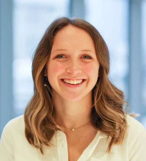 Professional headshot of a smiling woman with brown wavy hair wearing a cream-colored blouse in a modern office setting