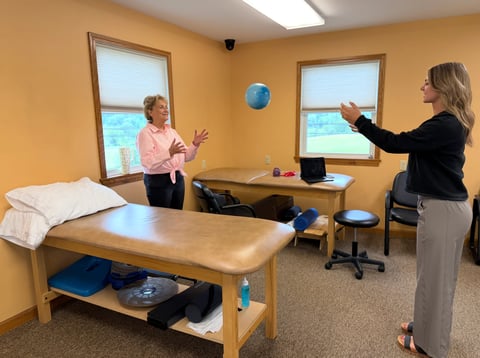 Two healthcare professionals in a simulated patient room with massage table and medical equipment, one demonstrating technique while the other observes by the window